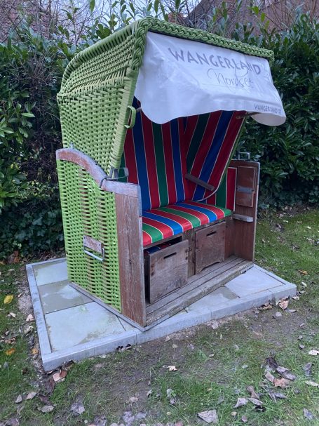 Bunte Strandkorb mit grünem Korbgeflecht und gestreiftem Sitzpolster auf einer Fläche.