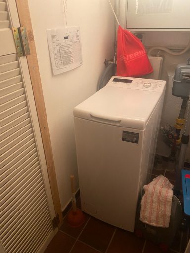 Waschmaschine in einer kleinen Nische, umgeben von Rohrleitungen und einer roten Tasche.