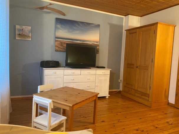 Heller Wohnraum mit Holzfußboden, TV, Kommode und einem kleinen Esstisch mit Stühlen.