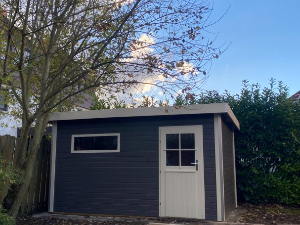 Gartenhaus mit grauer Fassade, weißer Tür und angrenzendem Baum vor blauem Himmel.