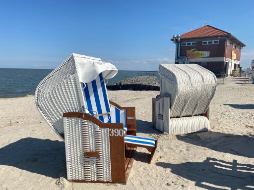 Zwei Strandkörbe auf sandigem Strand, im Hintergrund ein Gebäude und ein blauer Himmel.